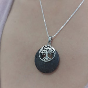 Può includere: Una collana con catena d'argento con un ciondolo rotondo che presenta un design ad albero della vita. Il ciondolo è realizzato in roccia lavica marrone scuro con un'incisione ad albero della vita argentata.