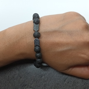 Puede incluir: Un brazalete de piedra de lava negra con cuentas de flecha plateadas.