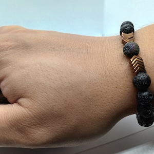 Peut inclure: Un bracelet en pierre de lave noire avec des perles en forme de flèche dorées. Le bracelet est porté au poignet.