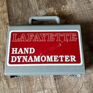 Könnte beinhalten: Ein grauer Kunststoffkoffer mit einem roten Etikett, auf dem "LAFAYETTE HAND DYNAMOMETER" steht, und einem kleineren Etikett mit der Aufschrift "CLICK CASE".