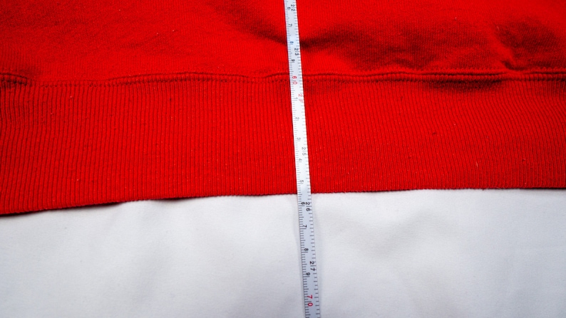 May include: A red sweatshirt with a ribbed hem. A white measuring tape is placed next to the hem, showing a measurement of 28 inches.