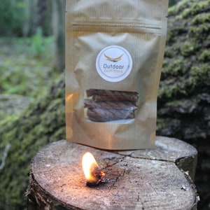 May include: A brown paper bag with a white label that says "Outdoor Outfitters" and a logo of a stylized arrow. The bag contains a bundle of brown fire starters. A fire starter is burning on a tree stump.