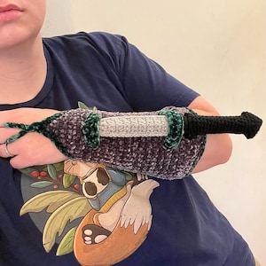 May include: A crocheted sword with a black handle, gray blade, and purple and green hilt. The sword is designed to be worn on the forearm. The design is handmade and features a textured, plush appearance.