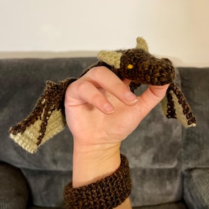 May include: A dark brown and light green crocheted dragon puppet featuring a textured body, pointed ears, and small yellow eyes. The dragon is attached to a matching dark brown crocheted wristband, creating a wearable accessory for the hand.