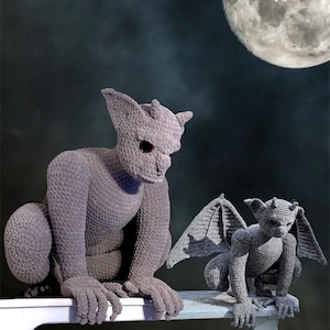 May include: Two gray crocheted gargoyle figurines. The larger gargoyle is sitting on a white surface. The smaller gargoyle is sitting on a gray surface. Both gargoyles have wings.