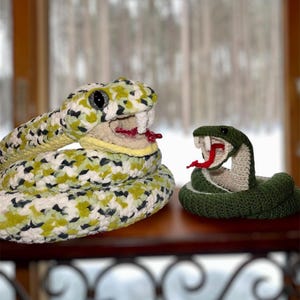 Op de afbeelding: Twee gehaakte slangen speelgoed, een groen en een met een groen, wit en geel patroon. De slangen zijn geposeerd met hun mond open, alsof ze op het punt staan ​​aan te vallen.