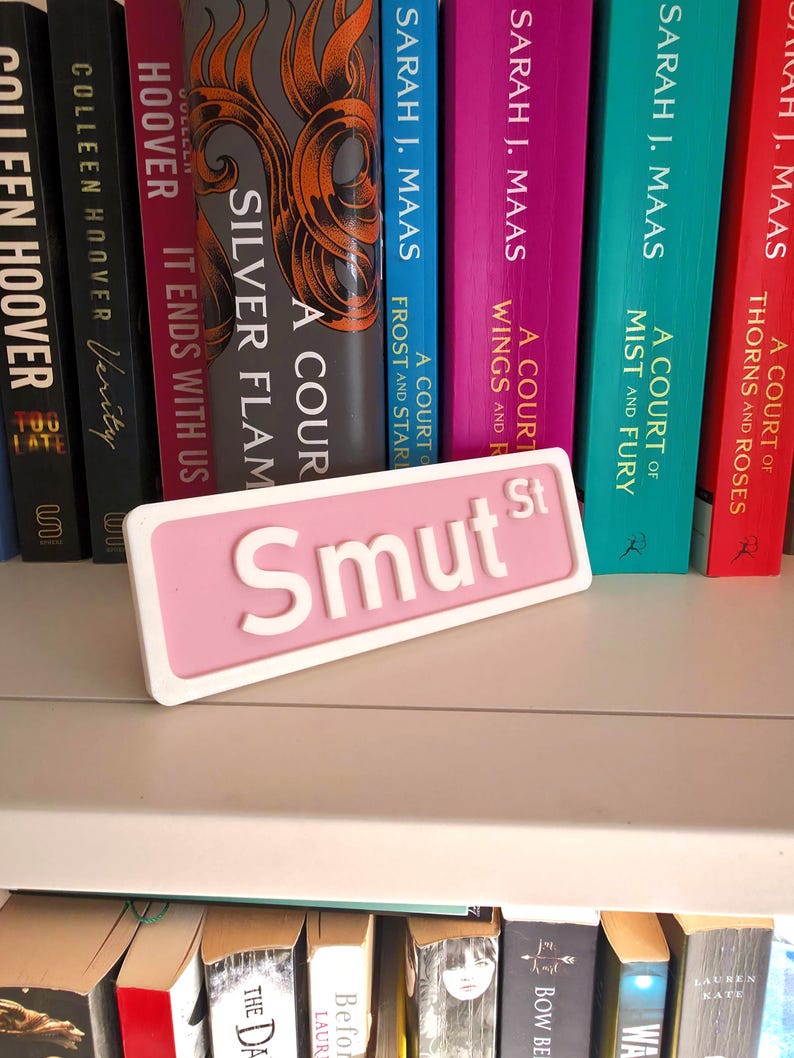 May include: A pink and white decorative sign that reads "Smut St" in white, placed in front of a bookshelf filled with colorful books. The sign is rectangular with rounded corners. The books have titles like "A Court of Thorns and Roses".