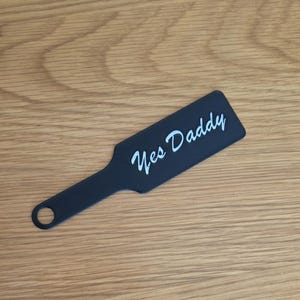 Puede incluir: Una paleta negra con un mango y un agujero en el extremo. Las palabras "Yes Daddy" están escritas en escritura cursiva blanca en la superficie de la paleta. La paleta está sobre una superficie de madera.