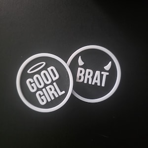 Puede incluir: Dos círculos negros con bordes y texto blancos. Uno dice "GOOD GIRL" con un halo, el otro "BRAT" con cuernos de diablo. El fondo es negro.