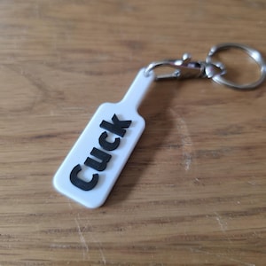 May include: White keychain with the word "Cuck" in black lettering. The keychain has a silver clasp and ring. The background is a wooden surface.