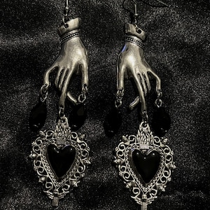 Puede incluir: Un par de pendientes de plata con una mano sosteniendo un corazón negro. El corazón está rodeado de un diseño de filigrana de plata. Los pendientes son colgantes y tienen cuentas negras.