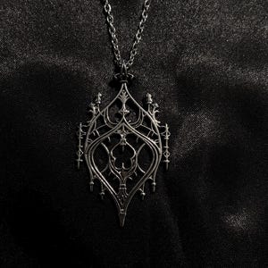 May include: Silver pendant necklace with a gothic-style design. The pendant has detailed filigree and a teardrop shape, hanging from a fine chain. The necklace is shown against a dark, textured backdrop.