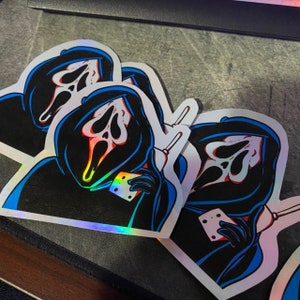 May include: Holographic stickers featuring the Ghostface character from the Scream movie franchise. The stickers depict Ghostface holding a phone, with a black robe, white mask, and blue outline. The stickers have a rainbow holographic effect.