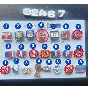 May include: A collection of square and shaped metal charms arranged on a white display card. The charms feature various colorful graphics, letters, and symbols related to the FIFA World Cup 26, with some marked with red SOLD OUT stamps.