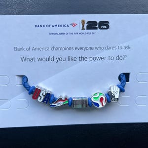 May include: A blue braided cord bracelet featuring five metal charms: a red, white, and blue USA charm, a soccer cleat, a rectangular block, a colorful soccer ball, and a goalkeeper glove with the number 26, displayed on a Bank of America card.
