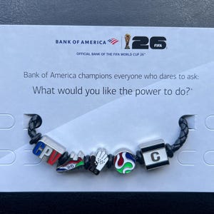 May include: A black braided bracelet featuring five metal charms: a cube with 'CPV', a soccer cleat, a goalie glove, a colorful soccer ball, and a cube with the letter 'C'. The bracelet is attached to a white card with Bank of America and FIFA World Cup 26 branding.