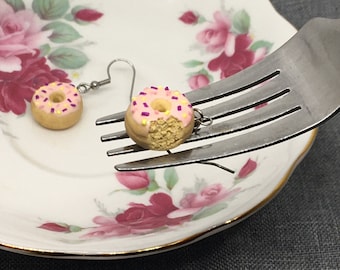 May include: A pair of pink and white donut earrings with purple sprinkles. The earrings are hanging from a fork.