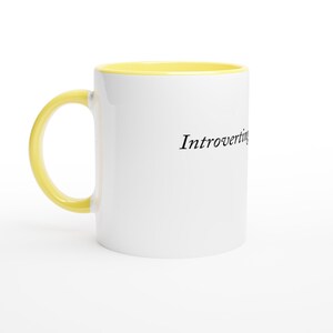 May include: White ceramic mug with a yellow interior and handle. The mug features the word "Introverti" in black script on the side. The mug is set against a white background.