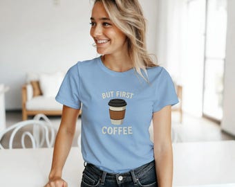 May include: Light blue t-shirt with the text "BUT FIRST COFFEE" and a graphic of a coffee cup. The shirt is made of a soft material and is suitable for casual wear. The shirt is paired with dark blue jeans.