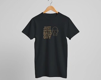 May include: Black t-shirt with the text "JUST ANOTHER BALD GUY" and a line drawing of a bald head in a light brown color. The t-shirt is hanging on a wooden hanger.