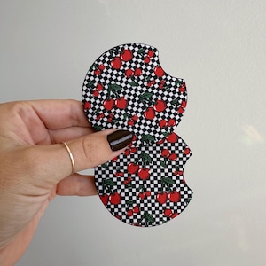 May include: Two car coasters with a black and white checkered pattern and red cherries. The coasters have a curved cutout for easy removal from a car cup holder. The cherries have green stems and leaves.