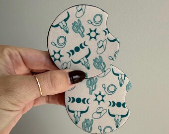 Puede incluir: Dos posavasos para coche de color blanquecino con un estampado de temática occidental en color verde azulado. El estampado incluye cactus, sombreros de vaquero, insignias de sheriff, calaveras de toro y fases lunares. Los posavasos son redondos con un recorte curvo.