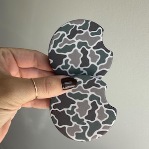 May include: Two round car coasters with a camouflage pattern in shades of gray, green, and white. Each coaster has a semi-circular cutout on one side. The coasters are held up by a hand.