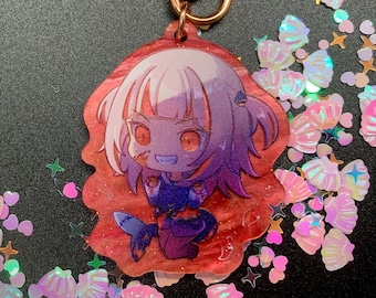 Könnte beinhalten: Ein pinkfarbener und violetter Anime-Charakter-Schlüsselanhänger mit einer goldenen Kette. Der Charakter ist ein Mädchen mit langen pinkfarbenen Haaren und roten Augen. Sie trägt ein blaues und violettes Outfit und hat einen Fischschwanz. Der Schlüsselanhänger ist von schimmerndem Glitzer umgeben.