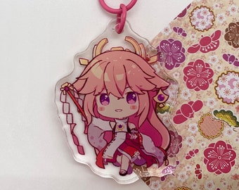 Może przedstawiać: Brelok z postacią anime w kolorze różowym z różowym metalowym zapięciem. Postać to młoda kobieta z długimi różowymi włosami, ubrana w białą i czerwoną kimonę. Tło to różowy i biały wzór kwiatowy.