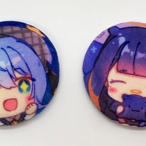 May include: Two round plush pins featuring anime characters. The first pin has a blue-haired girl with green eyes and a pink blush. The second pin has a purple-haired girl with yellow eyes and a pink blush. Both pins have a dark blue background with stars.