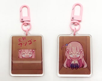 May include: A pink and white acrylic keychain featuring a cartoon character with pink hair and a blue bow. The character is sitting inside a cardboard box with the Japanese text "完成 マフー" printed on the box.