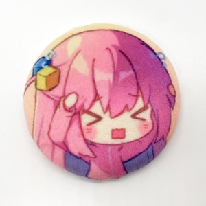 Pode incluir: Um botão redondo rosa e roxo com um personagem de desenho animado com cabelo rosa e uma expressão surpresa. O personagem está vestindo uma jaqueta roxa e azul.