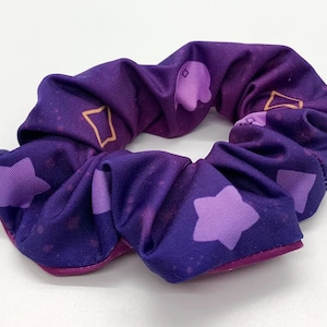 May include: Purple scrunchie with a galaxy print featuring stars and geometric shapes in shades of purple and gold.