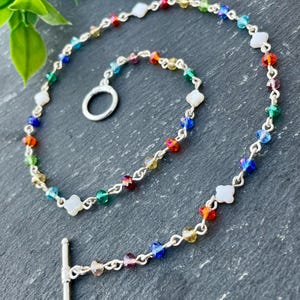 May include: A colorful beaded bracelet with a silver chain and clasp. The bracelet features a variety of rainbow-colored beads and white clover-shaped accents. The toggle clasp is silver. The bracelet is displayed on a dark gray surface.