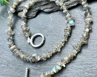 May include: A necklace made of small, irregularly shaped, gray-toned stones with flashes of blue and green. The necklace has a silver-colored toggle clasp. The stones are strung together to form a delicate, natural-looking piece of jewelry.