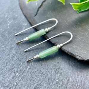 Op de afbeelding: Een paar zilveren oorbellen met een groene cilindrische kraal en kleine zilveren kralen. De oorbellen hebben een gebogen haakontwerp en een plat, rechthoekig uiteinde. De oorbellen worden gepresenteerd op een donkergrijze ondergrond.