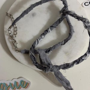 Puede incluir: Un collar de tela gris con un cierre de cadena plateada. El collar está hecho de tela retorcida con un borde deshilachado. La tela tiene un sutil patrón en blanco y negro. El collar está anudado en el centro.