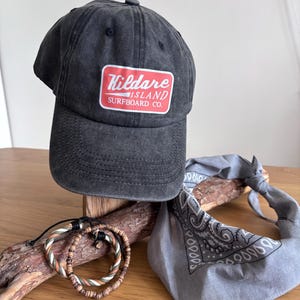 May include: A charcoal grey baseball cap with a rectangular patch featuring the red and white text Kildare Island Surfboard Co. Two beaded bracelets with green, white, and brown tones and a grey paisley patterned bandana are also present.