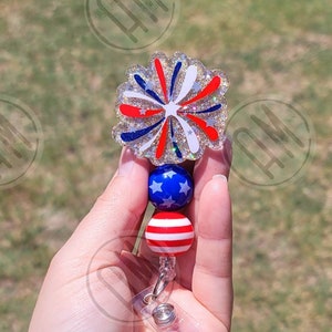 May include: A red, white, and blue glitter firework charm with a star-spangled bead and a red and white striped bead. The charm is attached to a clear retractable badge holder.