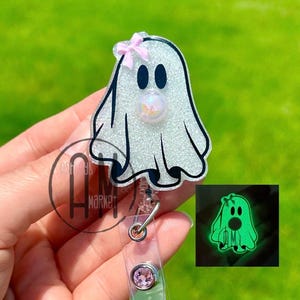 May include: A glittery white ghost-shaped badge reel with a pink bow and black eyes. The badge reel has a clear plastic strap and a pink gem. A smaller image shows the ghost glowing green.