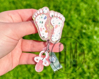 May include: Pink glitter baby feet charm with a pink pacifier charm attached to a retractable badge holder.