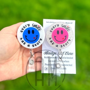May include: Two white glitter badge reels with a blue and pink smiley face design. The text on the reels reads "Every Smile Has A Story". The badge reels are attached to clear retractable badge holders. A white card with the text "Badge Reel Care" is visible in the background.