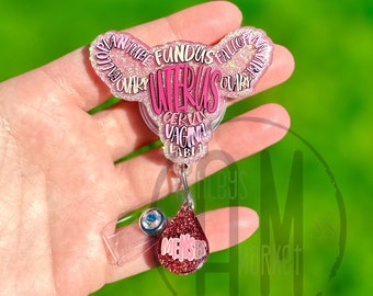 May include: Pink and white glitter resin badge holder shaped like a uterus with the word "uterus" and other anatomical terms written around it. The badge holder has a clear plastic badge clip and a pink glitter charm that says "menses".