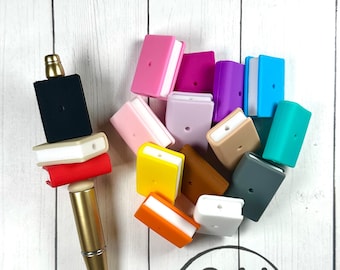 May include: A collection of colorful silicone book charms in various colors, including pink, purple, blue, yellow, orange, and brown. The charms are stacked on top of each other and are arranged in a circle. There is a gold pen in the image as well. The charms are labeled "Ashley's Market".
