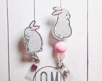 May include: Two bunny-themed retractable badge holders. Each badge holder features a glittery gray bunny with pink accents. One has a pink bead and clear beads. The badge reels have a clear plastic clip and a pink gem.