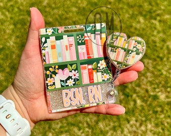 Può includere: Un porta badge verde e rosa glitter con un design a libreria e il testo "BSN. RN". Ha un ciondolo a forma di cuore abbinato con un design a libreria.