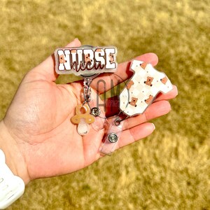 May include: A clear plastic badge reel with a brown and white patterned baby onesie charm, a pacifier charm, and a clear plastic badge holder with the text "NURSE" in white with a brown outline.