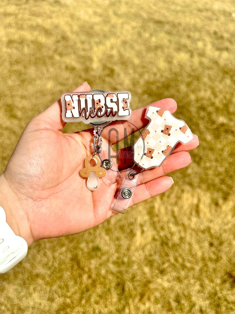 May include: A clear plastic badge reel with a brown and white patterned baby onesie charm, a pacifier charm, and a clear plastic badge holder with the text "NURSE" in white with a brown outline.