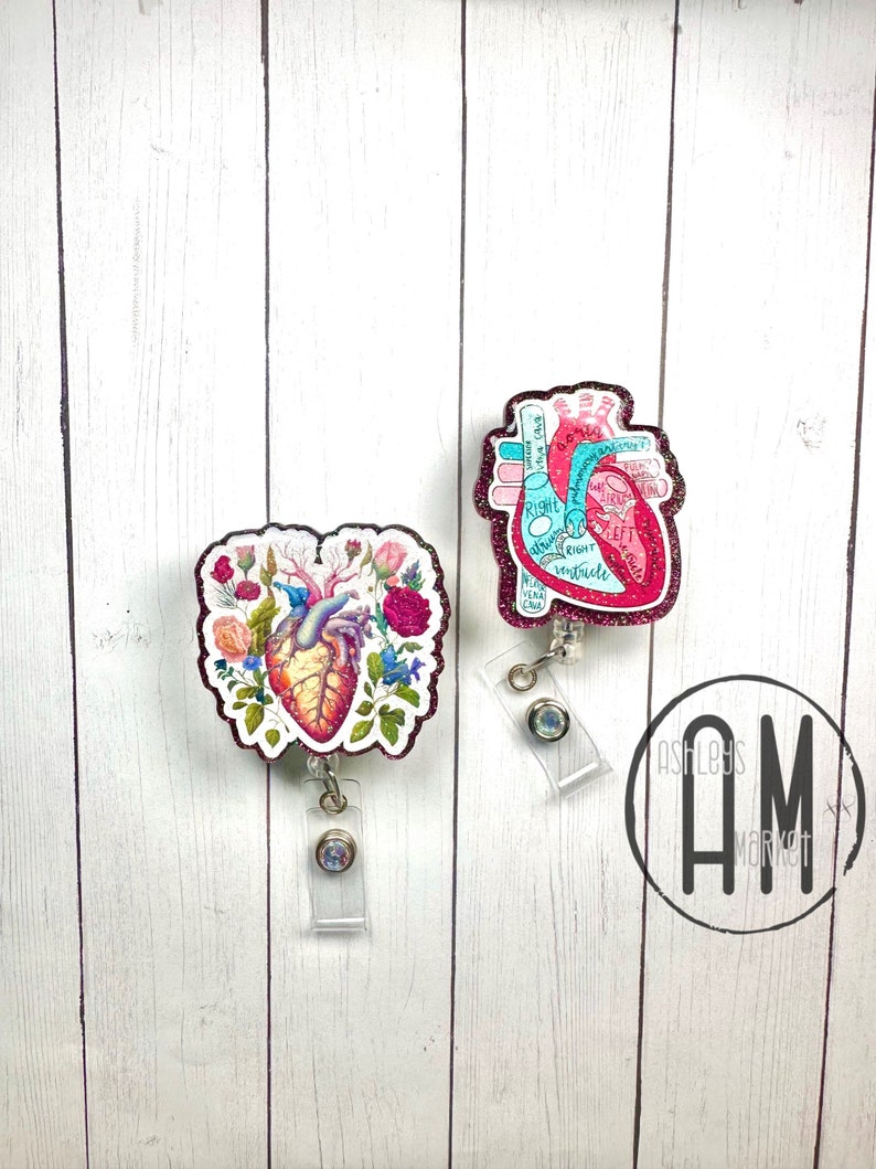 May include: Two heart-shaped badge reels with glitter accents. One reel features a detailed anatomical heart with labels, the other features a stylized heart with floral accents. Both reels have clear plastic clips and rhinestones.