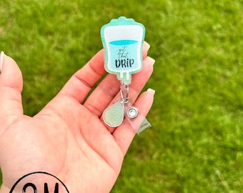 May include: A green glitter IV bag with the text "get that drip" on it. The IV bag is attached to a retractable badge reel with a clear plastic cover and a green glitter drop charm.
